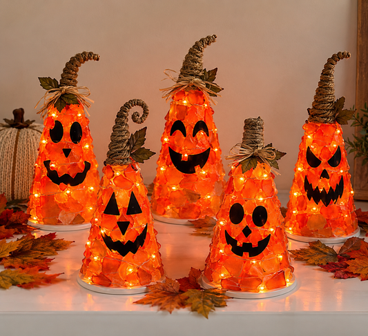 Five decorative Halloween jack-o'-lantern cone lanterns made from orange translucent leaves, each with a black face cutout, wrapped in warm white string lights, and topped with rustic brown raffia or twine hats. They are arranged on a white surface with scattered autumn leaves, with a neutral indoor background.